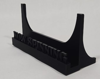 Può includere: Un espositore nero per dischi in vinile con la scritta "NOW SPINNING" a rilievo. Il supporto ha una base rettangolare e due supporti curvi. Il supporto è progettato per contenere un singolo disco.
