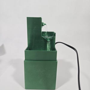 May include: A dark green tiered water fountain with a rectangular base and a vertical back panel. Water flows from the top tier down into a lower basin. A black electrical power cord extends from the side of the fountain structure.