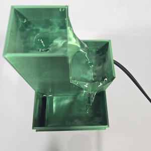 May include: A green tiered water fountain with a square structure. Water flows from the top level into a middle basin and then down into a bottom reservoir. A black power cord extends from the side of the middle section of the fountain.