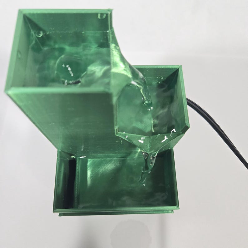 May include: A green tiered water fountain with a square structure. Water flows from the top level into a middle basin and then down into a bottom reservoir. A black power cord extends from the side of the middle section of the fountain.