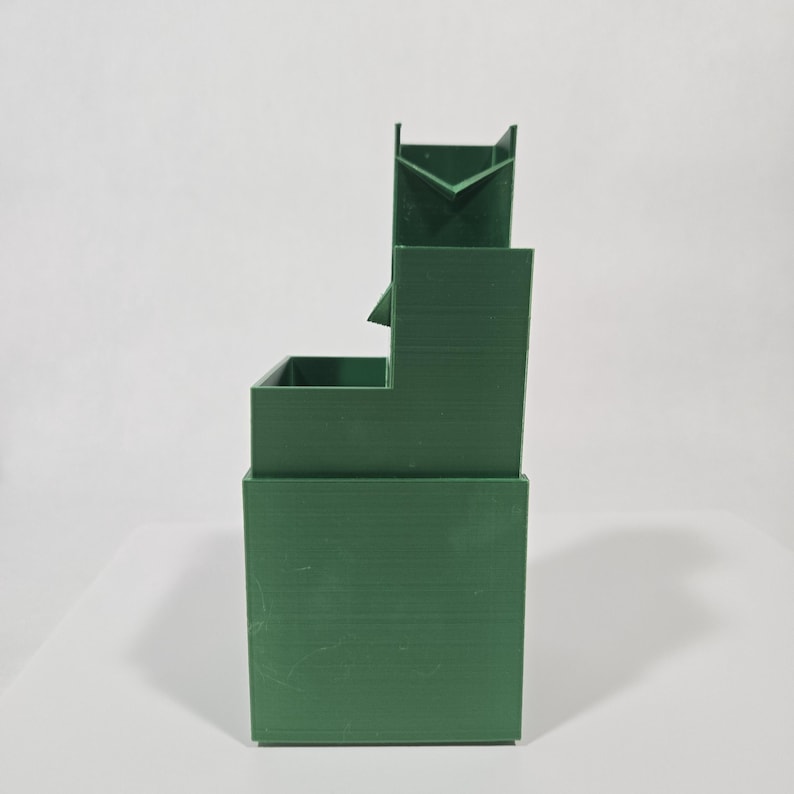May include: A dark green, multi-tiered desk organiser featuring a solid rectangular base, a mid-level square compartment, and a taller back section with an open, V-shaped cutout at the top. The object has a smooth, matte finish and structured lines.