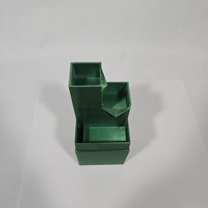 May include: A dark green plastic organiser with a tiered, stepped design featuring three distinct open compartments of varying heights. The structure has a square base and vertical walls, providing a functional storage solution for small items.
