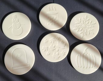 Può includere: Un set di sei sottobicchieri circolari bianchi con disegni in rilievo che includono un pupazzo di neve, una tazza fumante, un fiocco di neve, una lapide con il testo RIP, foglie d'acero e una zucca, disposti su una superficie scura.