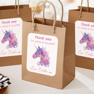 May include: Three brown paper gift bags with twisted paper handles. Each bag features a white label with a watercolour unicorn design and the text "Thank you for coming to my party!" and "Love Emilia xx".