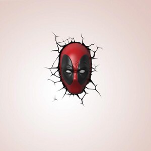 May include: A red and black Deadpool mask breaking through a cracked wall. The mask has black eye coverings and white eye details. The wall is a pale pink colour.