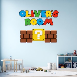 Può includere: Un adesivo murale con la scritta "OLIVER'S ROOM" in lettere colorate e a blocchi sopra un muro di mattoni marroni con un blocco giallo con un punto interrogativo. Il design ricorda un popolare videogioco.