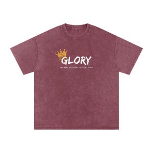 Könnte beinhalten: Ein kastanienbraunes Kurzarm-T-Shirt mit Vintage-Waschung. Auf der Brust befindet sich eine goldene Kronengrafik über dem weißen Schriftzug GLORY und dem kleineren weißen Text One step at a time, You'll get there. Das Shirt hat einen Rundhalsausschnitt.