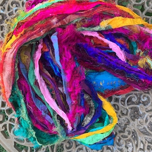 May include: A colourful bundle of silk ribbon in shades of pink, purple, blue, yellow, and green. The ribbons have a frayed texture and are arranged in a loose, layered pile. The image is taken from a high angle, showing the texture and colour variations.