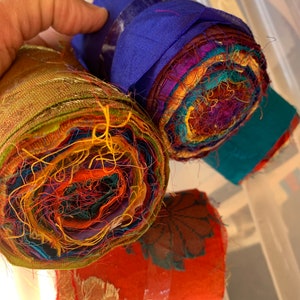 May include: Several rolls of colourful fabric, tightly wound and secured with clear tape. The rolls display a variety of vibrant colours, including blue, orange, green, and red. The edges of the fabric show a mix of textures and patterns, suggesting a craft or textile theme.