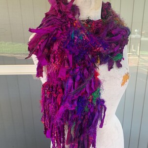 May include: A textured scarf made of various fabric strips in shades of magenta, purple, and violet, featuring frayed edges and mixed yarn textures. The scarf is draped around a mannequin, showcasing a layered, voluminous, and multi-dimensional appearance.