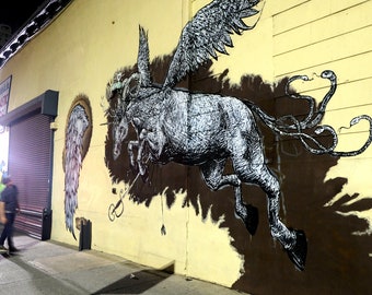 Puede incluir: Un mural de arte callejero en blanco y negro que representa un caballo alado con cola de serpiente, pintado en una pared amarilla. El caballo está en el aire, con las alas extendidas. La cola de serpiente está enroscada alrededor de las patas del caballo.