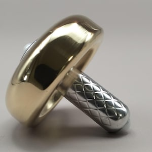 May include: A spinning top toy featuring a smooth, polished gold-toned circular disc attached to a silver-toned cylindrical stem. The stem has a textured, diamond-patterned knurled surface for grip, and a small white tip is visible at the base.