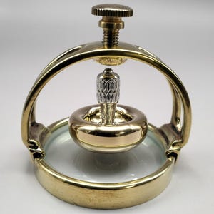 May include: A gold-toned metal gyroscope featuring a circular base and an arched frame with a top adjustment screw. The central spinning component has a polished gold-toned ring and a textured silver-toned vertical stem, all resting on a clear base.