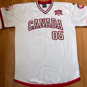 May include: White jersey with red and black detailing. The word "CANADA" is across the chest in red with a black outline, and the number "05" is below it. The jersey has a V-neck collar with red and black trim.