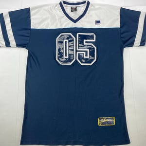 May include: Navy blue and white athletic jersey with the number "05" on the front. The jersey has white stripes on the sleeves and a white panel across the shoulders. A small logo is visible on the chest.