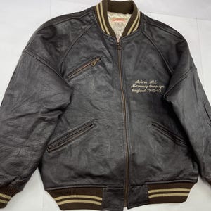 Pu&ograve; includere: Giacca bomber in pelle marrone scuro con chiusura a zip, due tasche frontali e colletto e polsini a coste. La giacca presenta un testo ricamato sul petto: "Avirex Ltd. Normandy Campaign England 1942-43."