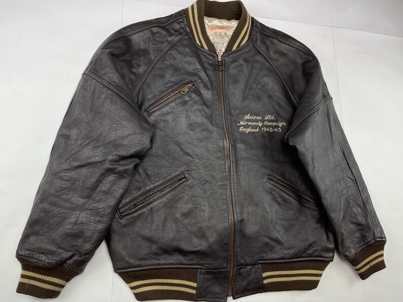 Pu&ograve; includere: Giacca bomber in pelle marrone scuro con chiusura a zip, due tasche frontali e colletto e polsini a coste. La giacca presenta un testo ricamato sul petto: "Avirex Ltd. Normandy Campaign England 1942-43."