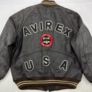 Pu&ograve; includere: Giacca in pelle marrone con la scritta "AVIREX" sulla schiena in lettere bianche. Sotto, una toppa circolare con le parole "AVIREX LTD" e "NEW YORK N.Y." Sono presenti anche le lettere "USA".