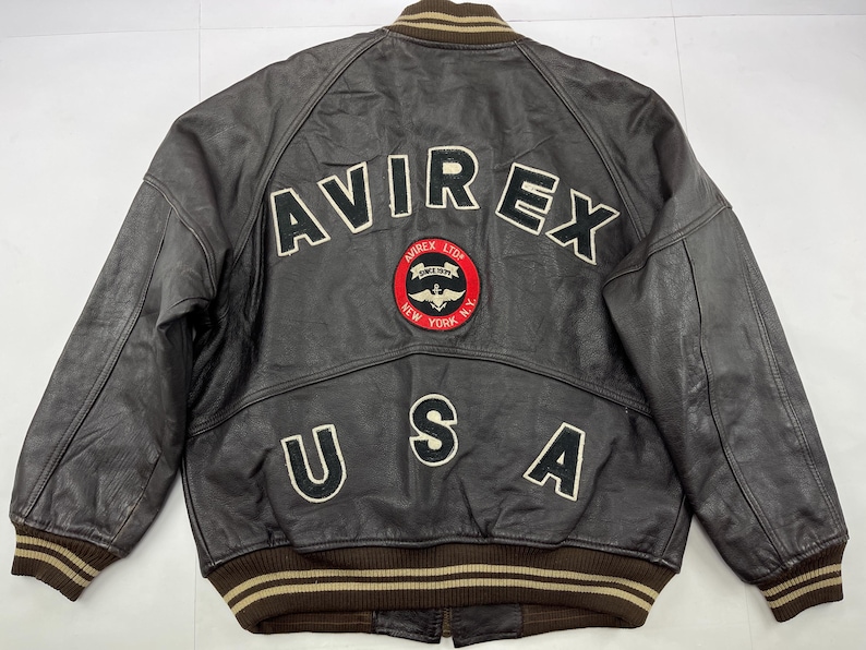 Pu&ograve; includere: Giacca in pelle marrone con la scritta "AVIREX" sulla schiena in lettere bianche. Sotto, una toppa circolare con le parole "AVIREX LTD" e "NEW YORK N.Y." Sono presenti anche le lettere "USA".