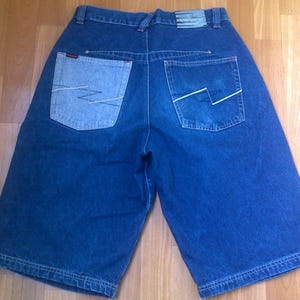 May include: Dark blue denim shorts featuring a high-waisted design with belt loops and a rectangular patch on the back waistband. The back includes two pockets, one in a lighter blue wash, both featuring white zigzag stitching and red accents.