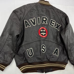 Pu&ograve; includere: Giacca bomber in pelle marrone con la scritta "AVIREX" in lettere bianche sul retro. Sotto, una toppa con "AVIREX LTD NEW YORK N.Y." e una grafica di un aereo. La parola "USA" &egrave; presente anche sul retro della giacca.