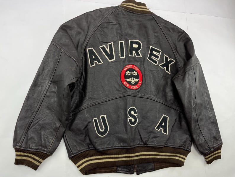 Pu&ograve; includere: Giacca bomber in pelle marrone con la scritta "AVIREX" in lettere bianche sul retro. Sotto, una toppa con "AVIREX LTD NEW YORK N.Y." e una grafica di un aereo. La parola "USA" &egrave; presente anche sul retro della giacca.