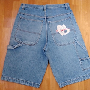 May include: Light blue denim shorts with a large white embroidered logo on the back pocket. The shorts have multiple pockets, including two large side pockets and two smaller pockets on the back.