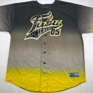 Peut inclure: Un maillot de baseball à boutons avec un dégradé de couleurs allant du jaune au gris et au noir. Le maillot affiche le texte "FUBU LEAGUE 05" dans une police stylisée. Un petit écusson est visible en bas.