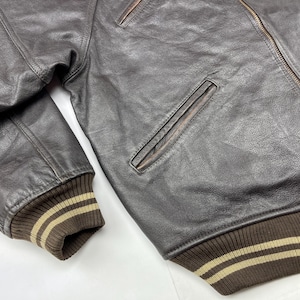 Pu&ograve; includere: Primo piano di un bomber in pelle marrone. La giacca presenta colletto, polsini e orlo a coste con strisce beige. Una tasca con cerniera &egrave; visibile sul petto. La pelle ha un aspetto invecchiato.