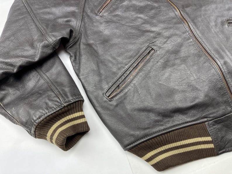 Pu&ograve; includere: Primo piano di un bomber in pelle marrone. La giacca presenta colletto, polsini e orlo a coste con strisce beige. Una tasca con cerniera &egrave; visibile sul petto. La pelle ha un aspetto invecchiato.