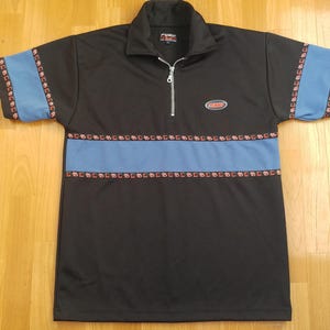 May include: A black short-sleeved polo shirt with a quarter-zip collar and a silver zip pull. The shirt features a horizontal light blue stripe across the chest and light blue sleeve cuffs, both bordered by a pattern of small red squares.