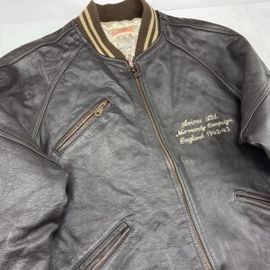 Pu&ograve; includere: Giacca bomber in pelle marrone scuro con chiusura a zip e due tasche frontali. La giacca ha un colletto a coste e il testo ricamato "Avirex Ltd Normandy Campaign England 1942-43".