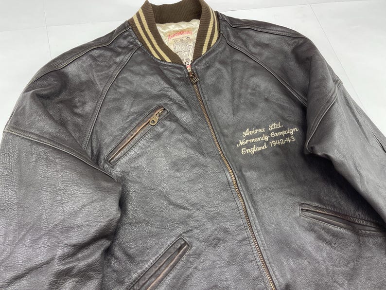 Pu&ograve; includere: Giacca bomber in pelle marrone scuro con chiusura a zip e due tasche frontali. La giacca ha un colletto a coste e il testo ricamato "Avirex Ltd Normandy Campaign England 1942-43".