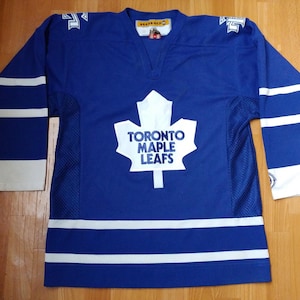 May include: Blue hockey jersey with white stripes on the sleeves and a white and blue Toronto Maple Leafs logo on the chest. The jersey has a V-neck and a mesh panel on the sides.
