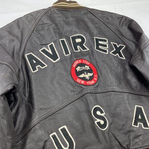 Pu&ograve; includere: Giacca di pelle marrone con la scritta "AVIREX" a grandi lettere bianche sulla schiena. Sotto, una toppa circolare con le parole "AVIREX LTD NEW YORK N.Y." e una grafica. Le lettere "USA" sono cucite sotto.