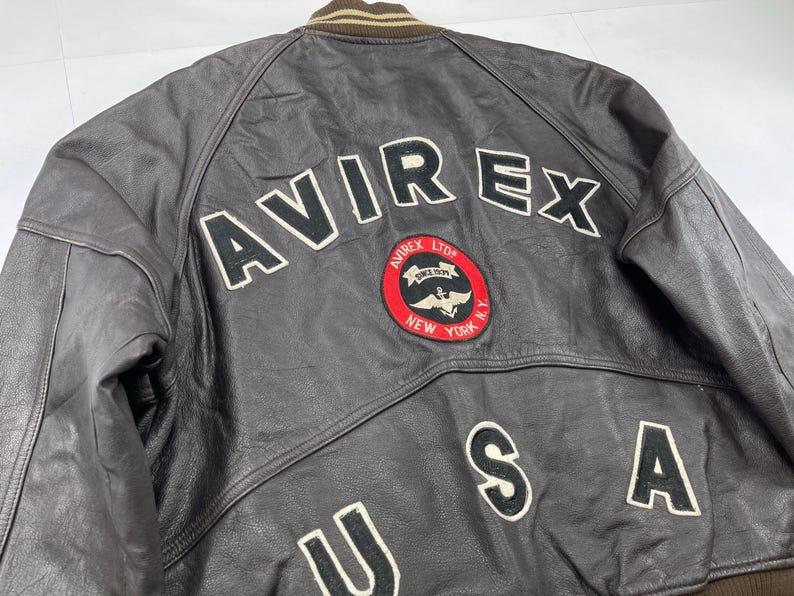 Pu&ograve; includere: Giacca di pelle marrone con la scritta "AVIREX" a grandi lettere bianche sulla schiena. Sotto, una toppa circolare con le parole "AVIREX LTD NEW YORK N.Y." e una grafica. Le lettere "USA" sono cucite sotto.