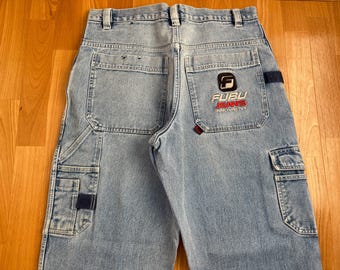 May include: Light blue denim jeans with multiple pockets and the "FUBU JEANS INNOVATIVE" logo embroidered on the back pocket. The jeans have a classic, relaxed fit and a faded wash. The jeans are laid flat on a wooden surface.