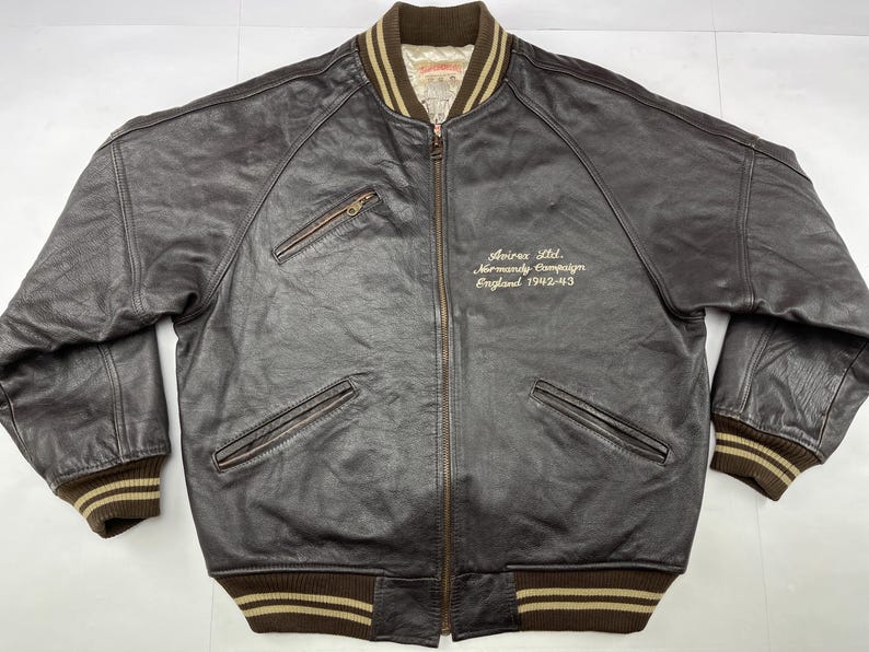 Pu&ograve; includere: Giacca bomber in pelle marrone con chiusura a zip, due tasche frontali e polsini e orlo a coste. La giacca presenta la scritta "Avirex Ltd. Normandy Campaign England 1942-43" ricamata sul petto.