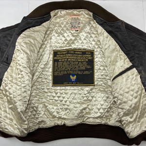 Pu&ograve; includere: Giacca bomber vintage in pelle marrone con fodera trapuntata color crema. La giacca presenta colletto e polsini a coste marroni. All'interno, un'etichetta mostra il testo "AVIREX USA" e "AUTHENTIC AVIREX LTD FLIGHT GARMENT".