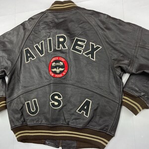 Pu&ograve; includere: Giacca bomber in pelle marrone con la scritta "AVIREX" sulla schiena in lettere bianche. Una toppa circolare con un logo e le parole "AVIREX LTD. NEW YORK N.Y." &egrave; centrata sotto le lettere. La parola "USA" &egrave; sotto.