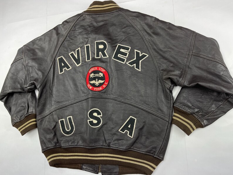 Pu&ograve; includere: Giacca bomber in pelle marrone con la scritta "AVIREX" sulla schiena in lettere bianche. Una toppa circolare con un logo e le parole "AVIREX LTD. NEW YORK N.Y." &egrave; centrata sotto le lettere. La parola "USA" &egrave; sotto.