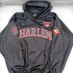 May include: A black hooded sweatshirt featuring the word "HARLEM" in red and white lettering. The sweatshirt has a "FUBU ATHLETICS 1992" patch and a red and white striped hem.