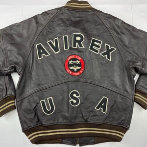 Pu&ograve; includere: Giacca in pelle marrone con la scritta "AVIREX" in lettere bianche sul retro. Sotto, una toppa circolare con "AVIREX LTD NEW YORK N.Y." e la scritta "USA". Il colletto e i polsini sono a coste marroni e beige.