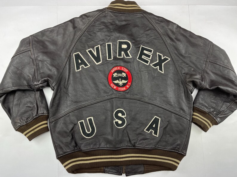 Pu&ograve; includere: Giacca in pelle marrone con la scritta "AVIREX" in lettere bianche sul retro. Sotto, una toppa circolare con "AVIREX LTD NEW YORK N.Y." e la scritta "USA". Il colletto e i polsini sono a coste marroni e beige.