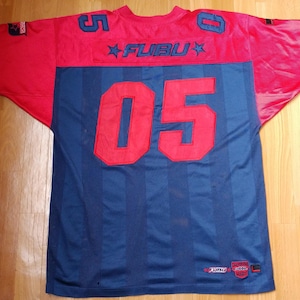 Può includere: Una maglia a maniche corte rossa e blu navy con la scritta FUBU sulla parte superiore della schiena. La maglia mostra il numero 05 in grande carattere rosso sul retro, con numeri 5 e 0 più piccoli sulle spalle. Presenta strisce verticali e toppe.