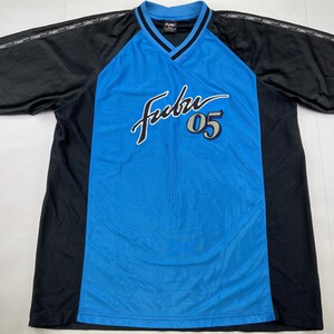 Può includere: Una maglia sportiva blu e nera con la scritta "Fubu" in corsivo bianco e il numero "05" in grigio. La maglia ha uno scollo a V e maniche corte. Le maniche e i pannelli laterali sono neri, mentre il davanti è blu.
