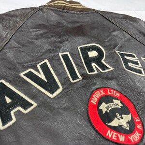 Pu&ograve; includere: Primo piano di una giacca di pelle marrone con la scritta "AVIREX" in grandi lettere nere bordate di bianco sul retro. &Egrave; visibile anche una toppa circolare con testo e logo.