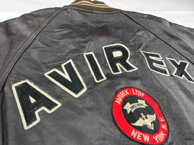 Pu&ograve; includere: Primo piano di una giacca di pelle marrone con la scritta "AVIREX" in grandi lettere nere bordate di bianco sul retro. &Egrave; visibile anche una toppa circolare con testo e logo.