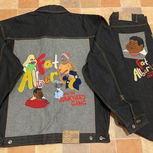 May include: A dark denim jacket and jeans set. The jacket features a grey panel on the back with embroidered characters from "Fat Albert and the Junkyard Gang." The jeans have a matching graphic on the back pocket.