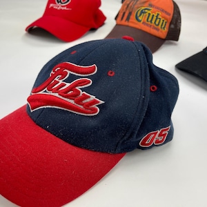 May include: A navy blue and red baseball cap with the word "Fubu" embroidered in white and red on the front. The cap has a red brim and a white "05" embroidered on the side.